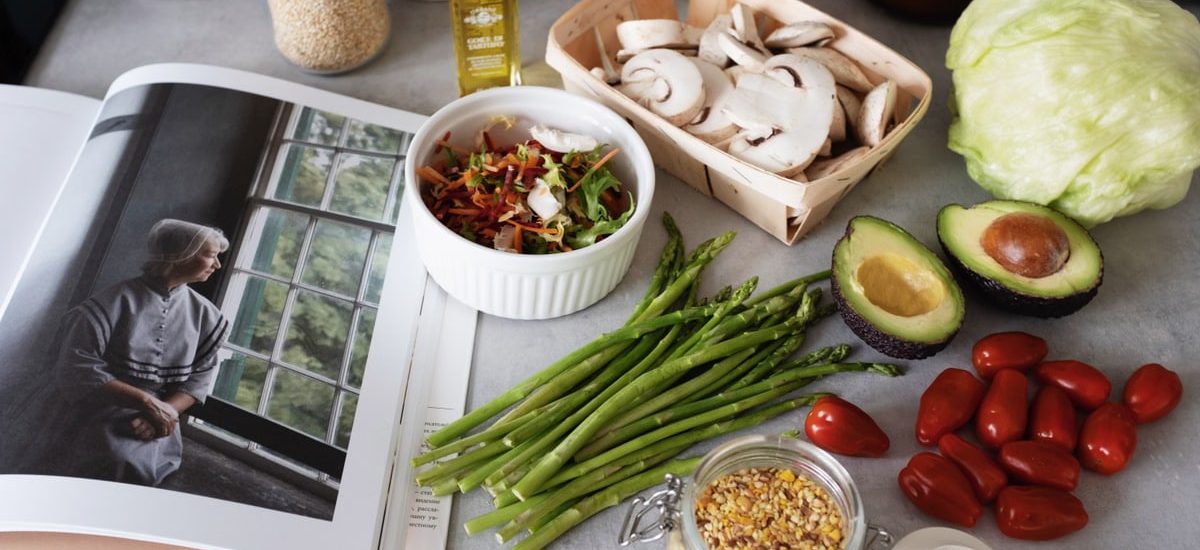 A selection of fresh vegetables and a cookbook in a kitchen setting, promoting healthy cooking.
